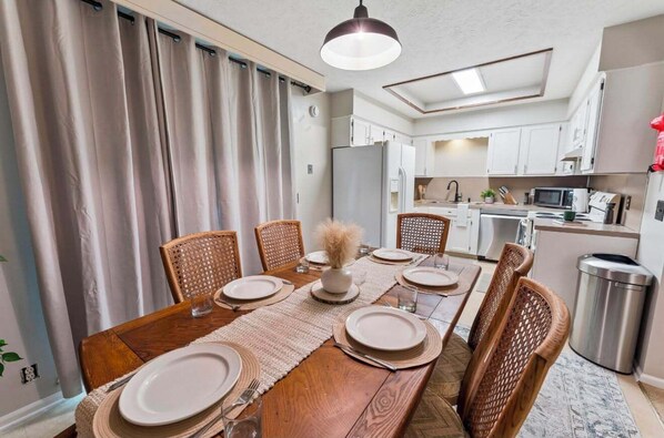 A dining area with a wooden table set for six, surrounded by wicker chairs, with a kitchen in the background.