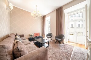 Un salon moderne avec un canapé, une table basse et deux fauteuils. On y trouve un lustre, une fenêtre avec des rideaux et une porte donnant sur l’extérieur.