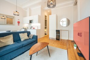 Ein modernes Wohnzimmer mit einem blauen Sofa, einem Holztisch und einer Küchenzeile mit weißen Schränken und einer Mikrowelle.