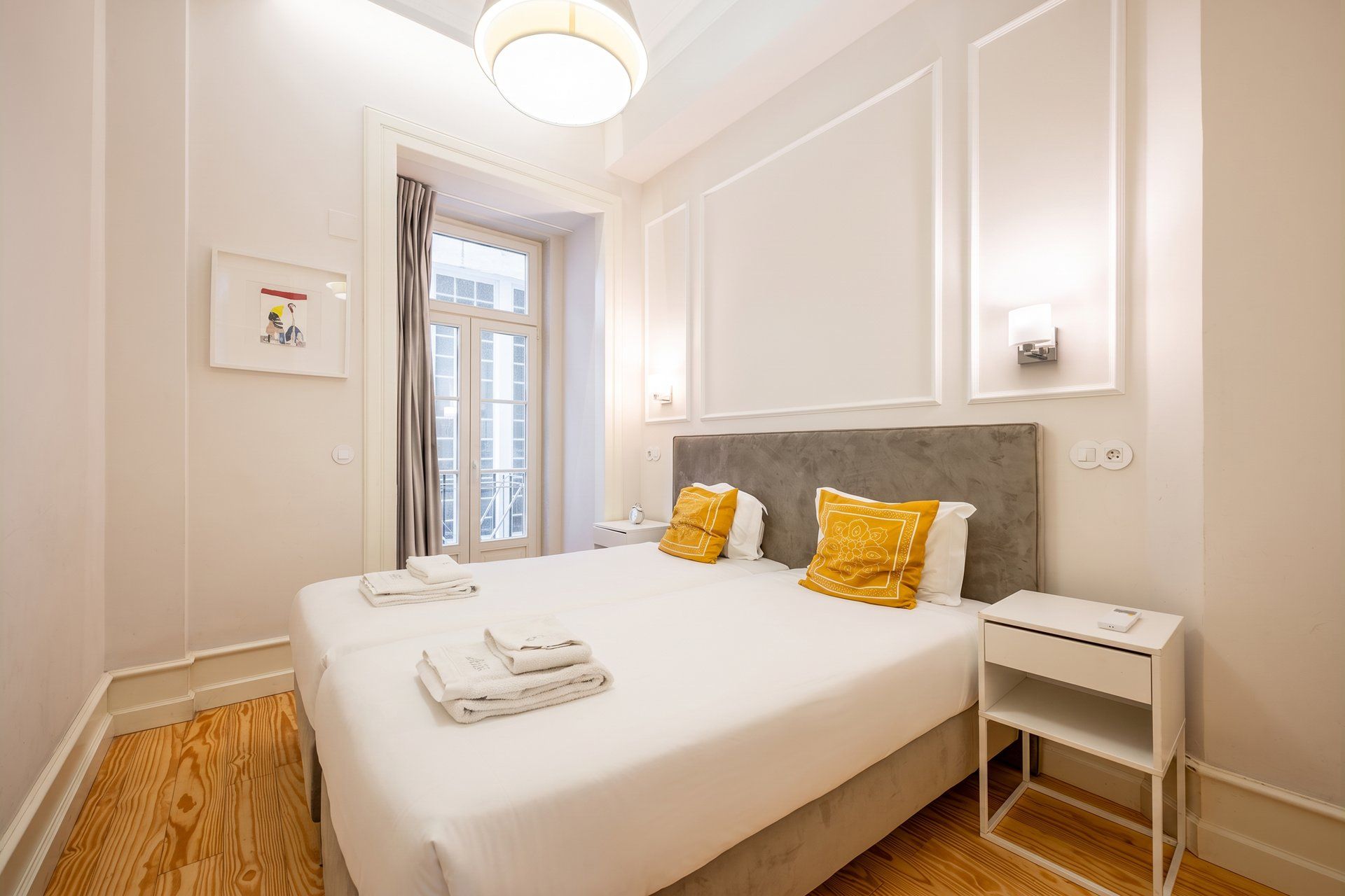 A hotel room with a large bed, a bedside table, a nightstand, a window with curtains, and a wall-mounted light fixture.