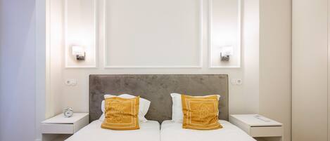 Cama doble con dos mesitas de noche, cada una con una lámpara, en una habitación con pared blanca lisa y cabecero sencillo.