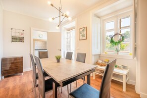 A dining area with a wooden table, chairs, a chandelier, a clock, and a window with a wreath. - GuestReady - Lemon House with Private Garden (Lisbon)