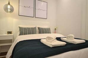 Un lit bien fait, avec du linge de lit blanc et deux serviettes enroulées, une table de chevet avec une lampe, et deux tableaux encadrés accrochés au mur.