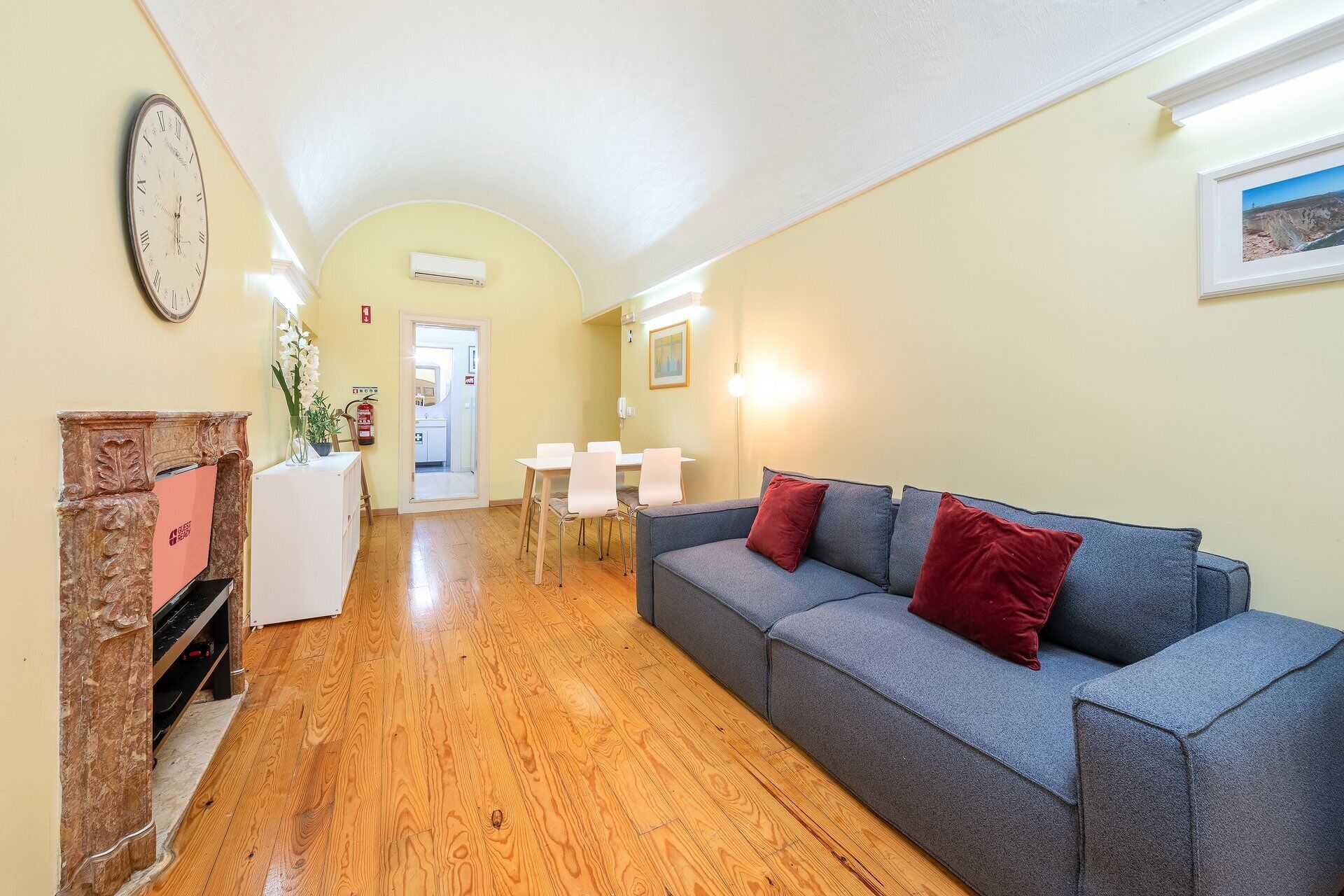 A hallway with a grey sofa, red cushions, a clock, and a framed picture on the wall.