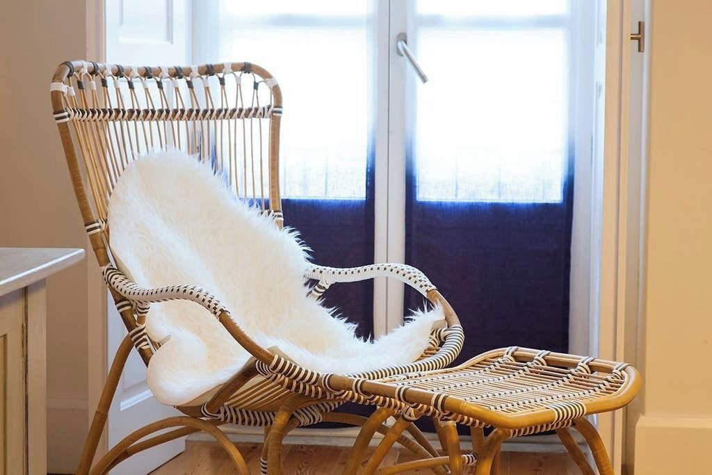 A wicker chair with a white furry seat and backrest, positioned near a window with a view of the outside.