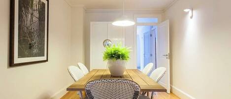 A dining area with a wooden table, white chairs, a potted plant, and a framed picture on the wall.