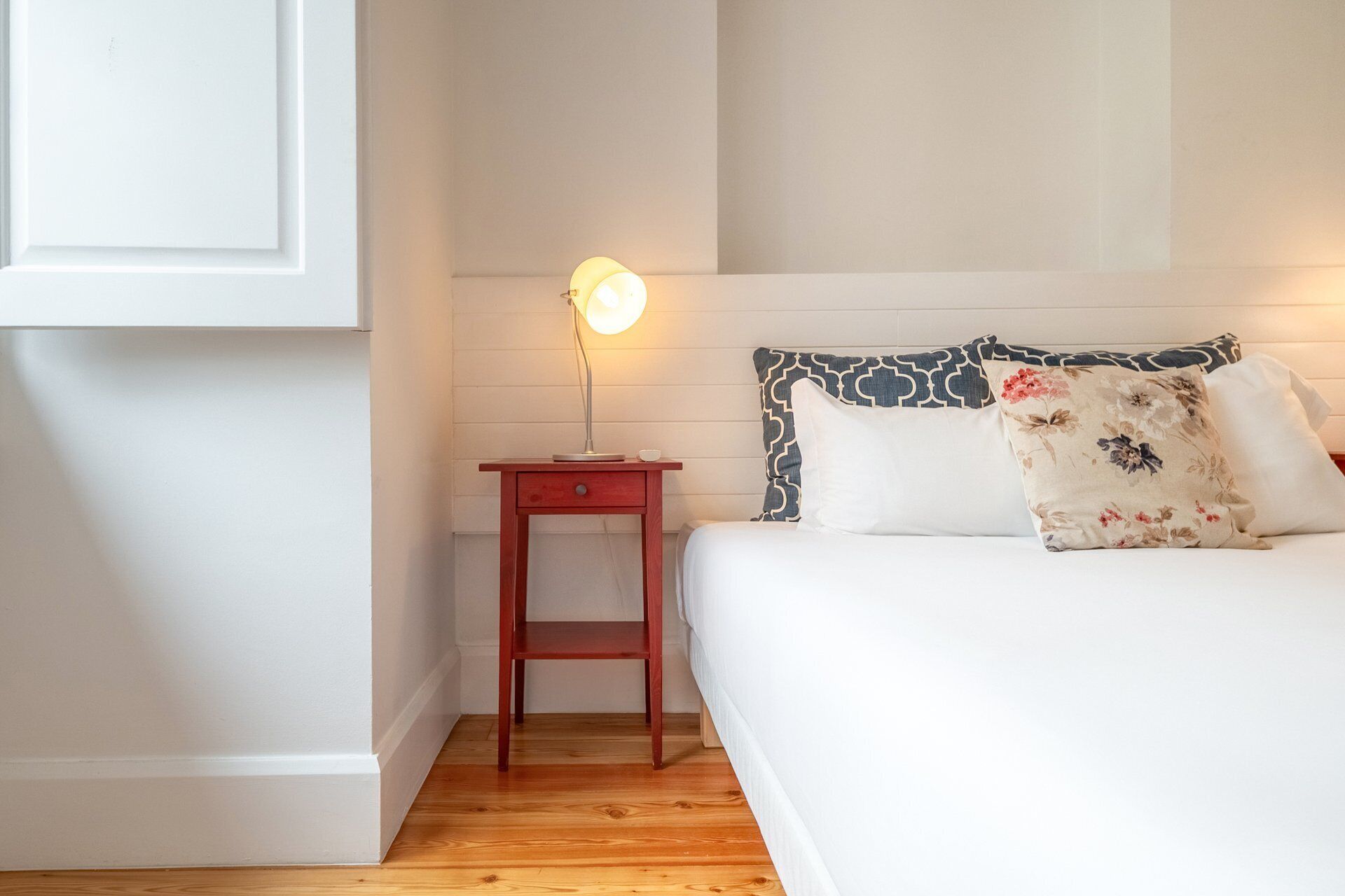A bedroom with a wooden floor, a bedside table with a lamp, a bed with patterned pillows, and a white wall.