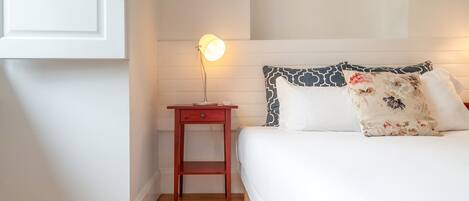 A bedroom with a wooden floor, a bedside table with a lamp, a bed with patterned pillows, and a white wall.