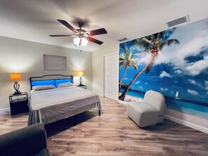 A bedroom with a bed, a ceiling fan, a nightstand with a lamp, a chair, and a wall mural of a beach scene. - West Melboburne Oasis, minutes to the beach! (Melbourne)