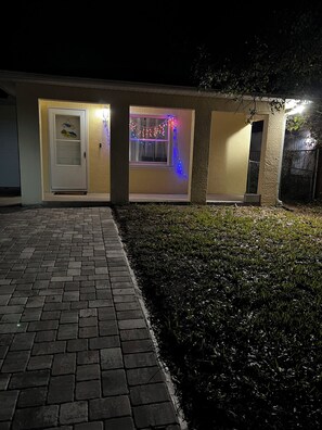 Een gebouw met een met baksteen bestrate oprit die naar een deur leidt, een klein raam met kleurrijke verlichting, en een gazon.