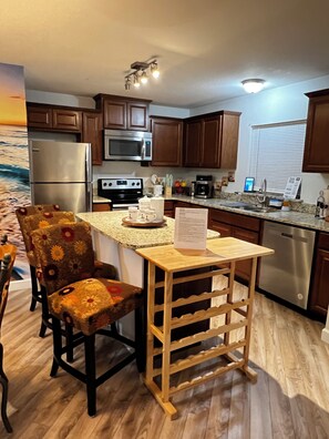 A kitchen with wooden cabinets, a stainless steel refrigerator, a microwave, a stove, and a wooden dining table with chairs. - West Melboburne Oasis, minutes to the beach! (Melbourne)