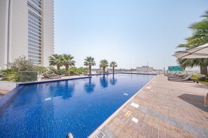 Ein Swimmingpool mit einer gefliesten Kante, umgeben von Palmen und Liegestühlen, mit einem Hochhaus im Hintergrund.
