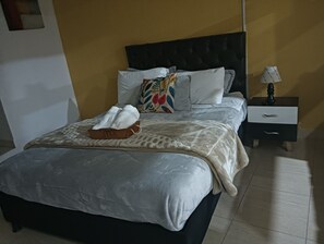 Un letto rifatto con un copriletto bianco e cuscini, un comodino con una lampada e un tavolino con un cassetto.