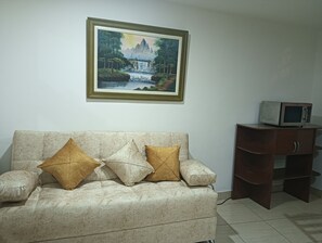 Ein Wohnzimmer mit einem beigefarbenen Sofa, zwei Kissen, einem gerahmten Gemälde einer Wasserfälle und einer Mikrowelle auf einem Holzkabinett.