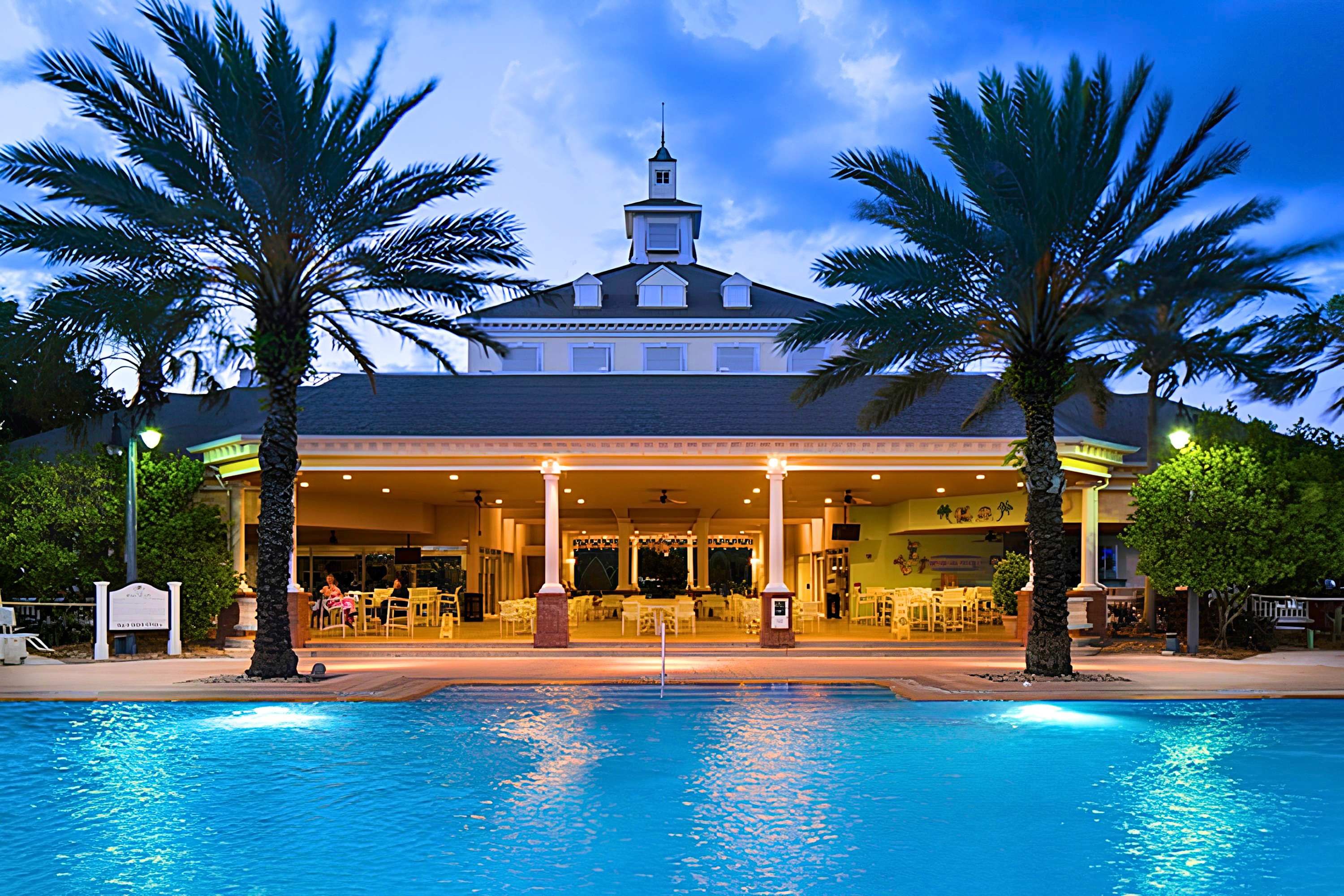 Un hotel con piscina, palmeras y una zona exterior cubierta con asientos.