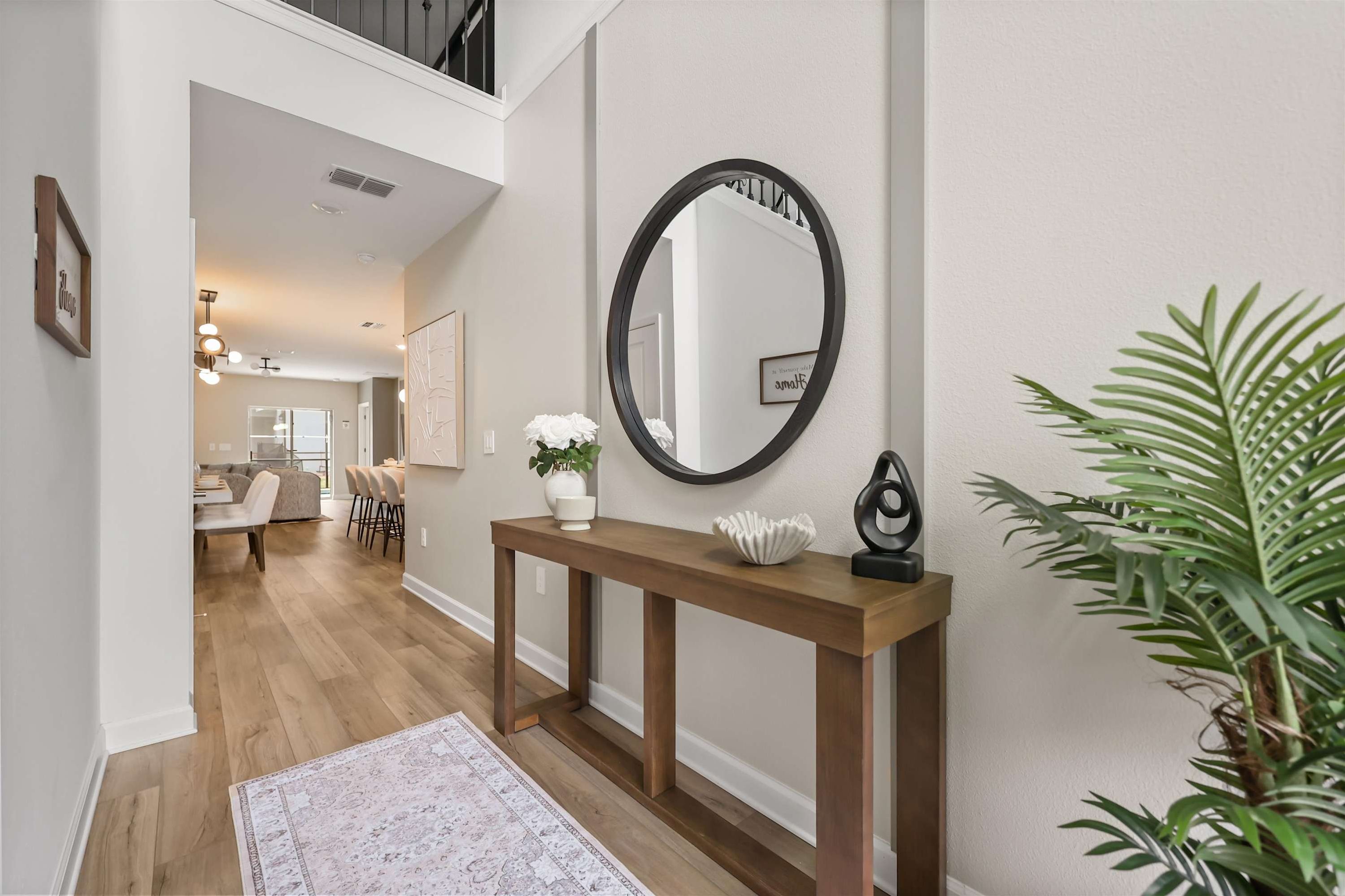 A hallway with a wooden console table, a round mirror, a potted plant, and a dining area in the background.