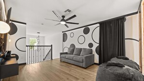 Una sala de estar moderna con un sofá gris, un sillón informal, un ventilador de techo y una escalera que lleva a un nivel superior.
