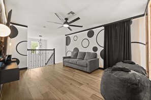 Una sala de estar moderna con un sofá gris, un sillón informal, un ventilador de techo y una escalera que lleva a un nivel superior.