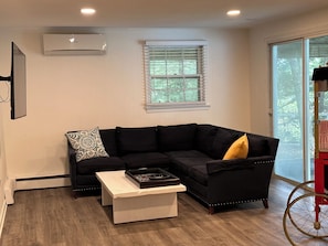 A living room with a black sectional sofa, a white coffee table, a window with blinds, and an air conditioning unit.