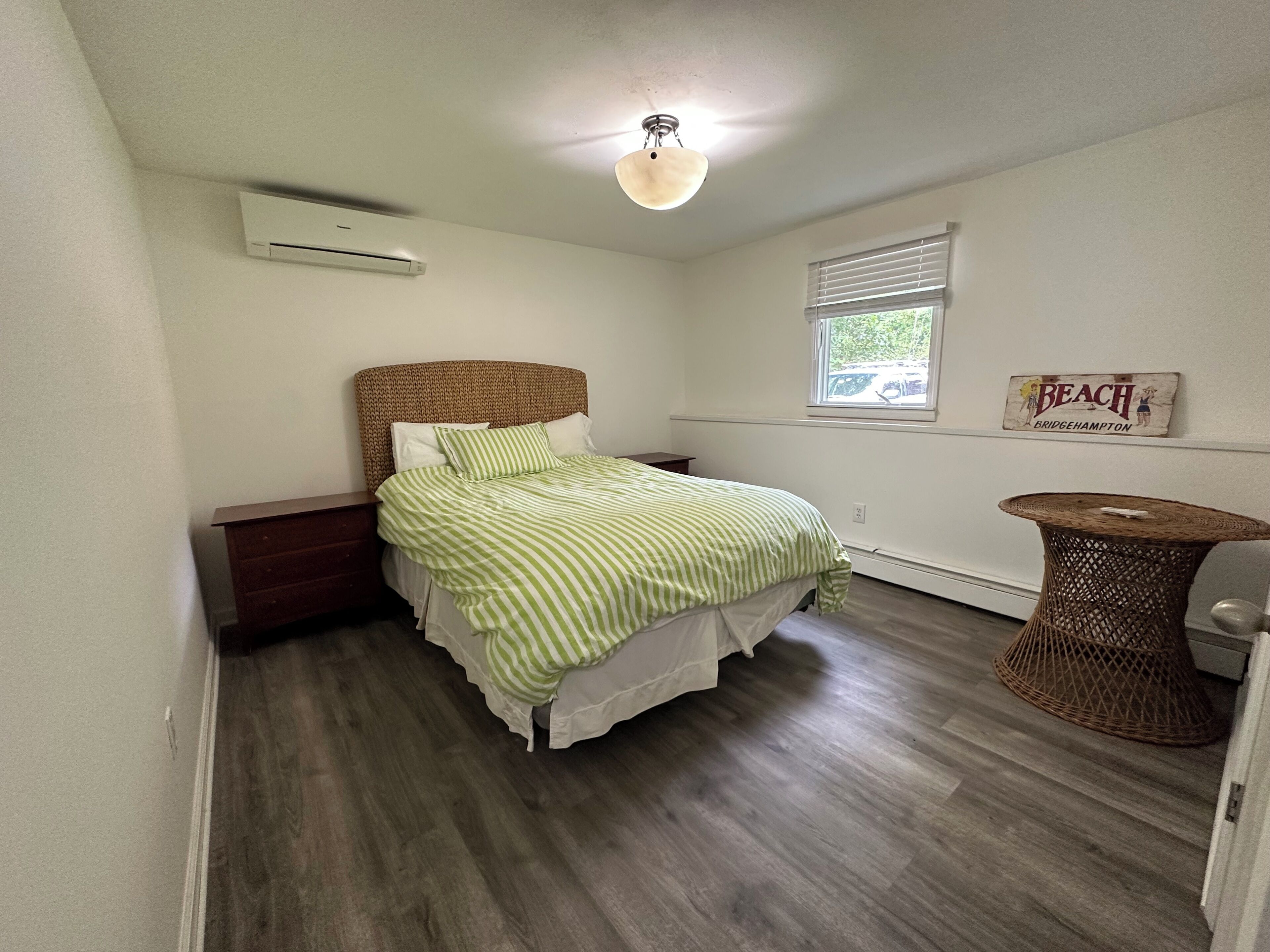 Ein Schlafzimmer mit Holzboden, einem Bett mit grün-weiß gestreifter Bettwäsche, einem Korbstuhl-Nachttisch, einer Wand-Klimaanlage und einem Fenster mit Jalousien.