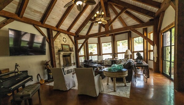 A spacious room with a high ceiling, wooden beams, a fireplace, a piano, and a large window.