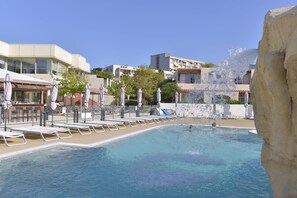 Una piscina con sillas de descanso y sombrillas, rodeada de edificios y un acantilado rocoso.
