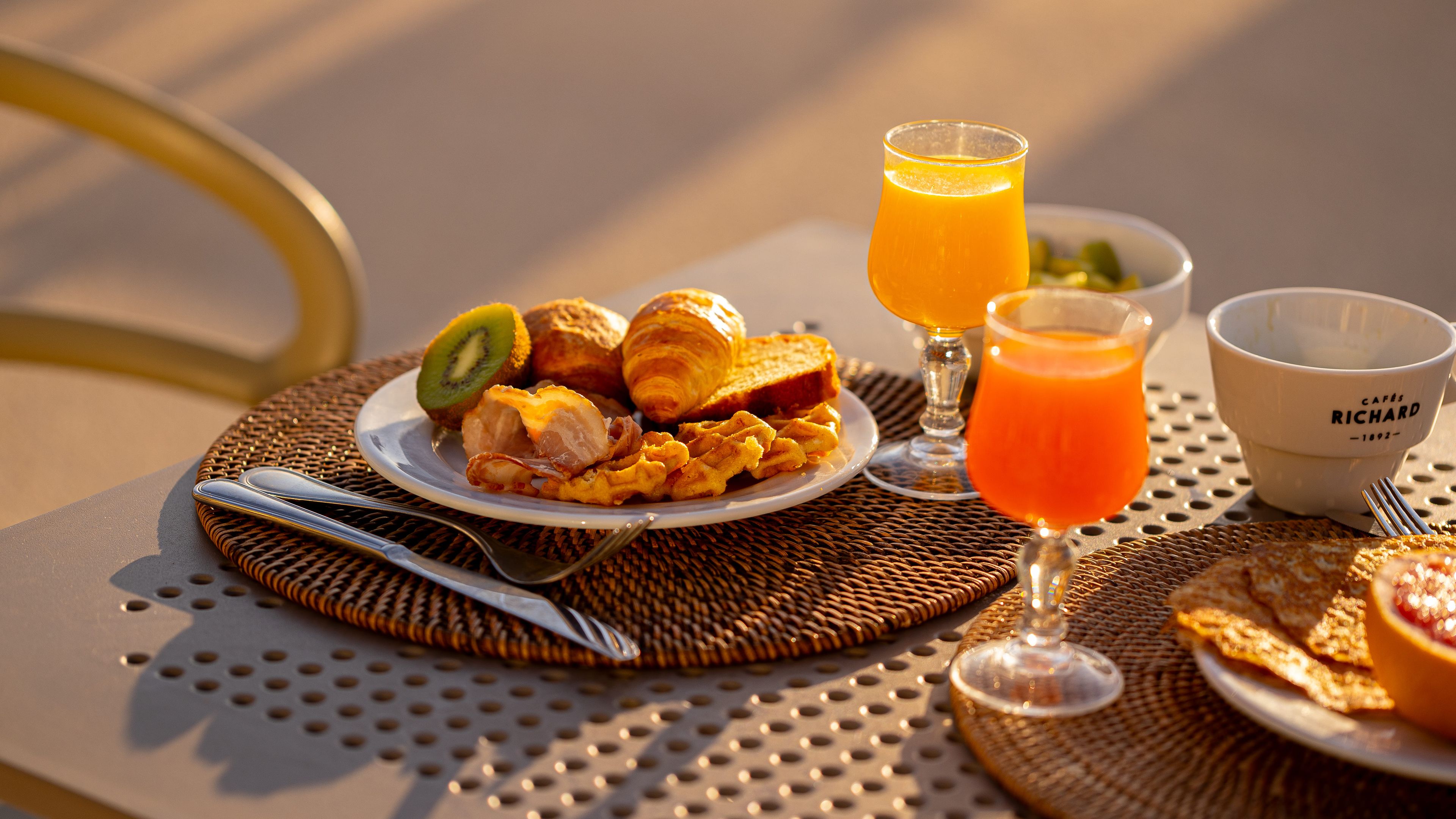 A breakfast setting with croissants, a kiwi slice, waffles, a glass of orange juice, and a cup labeled 'Cafés Richard.'