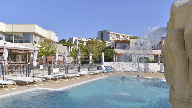 Una piscina con lettini e ombrelloni, circondata da edifici e una scogliera rocciosa.