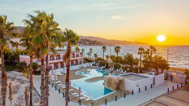 Um resort com piscina, palmeiras e vista para o oceano ao pôr do sol.