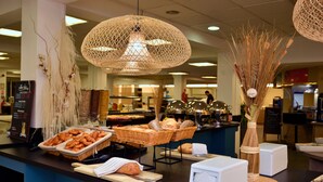 Buffet com diversos alimentos, incluindo pão e doces, em ambiente interno bem iluminado com luminárias pendentes.
