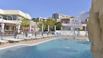 A swimming pool with lounge chairs and umbrellas, surrounded by buildings and a rocky cliff.