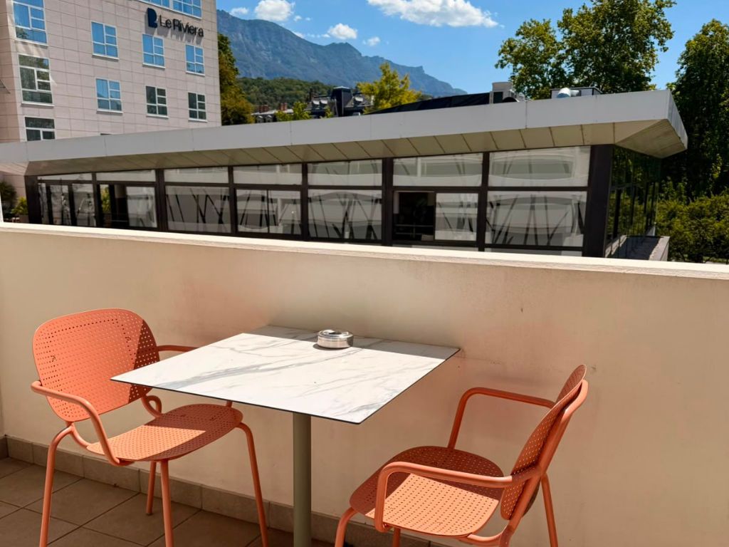 Een terras met twee oranje stoelen en een tafel, met uitzicht op een gebouw met een berg op de achtergrond.