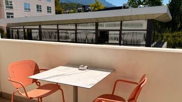Een terras met twee oranje stoelen en een tafel, met uitzicht op een gebouw met een berg op de achtergrond.