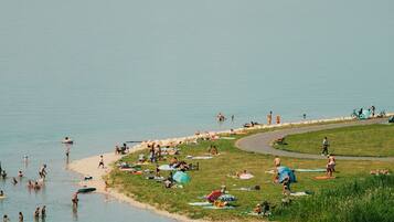 A lakeside area with people swimming and sunbathing, a grassy area with tents, and a boat in the water.