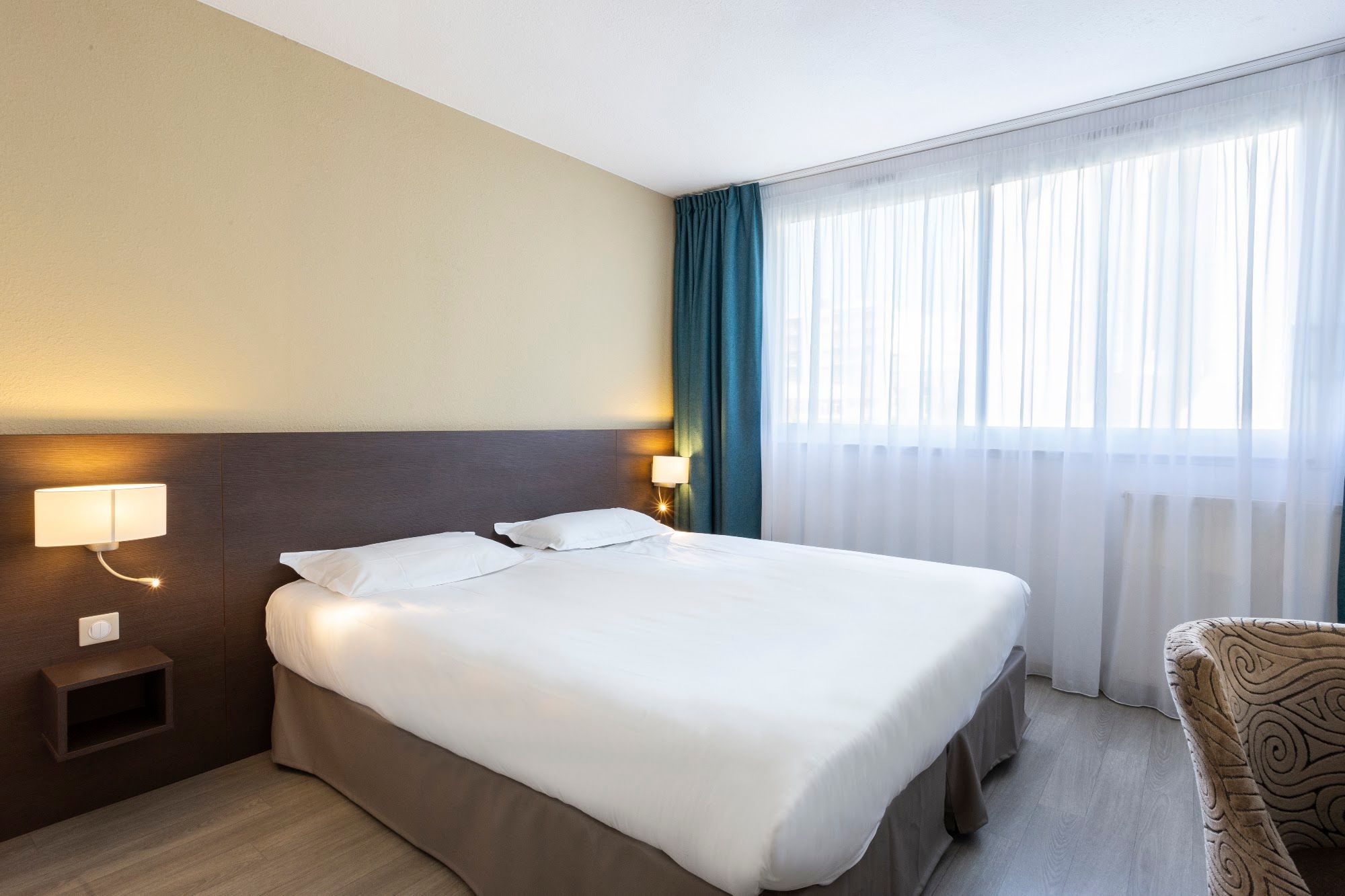 Una habitación de hotel con cama, lámparas de noche, una ventana con cortinas y una silla.