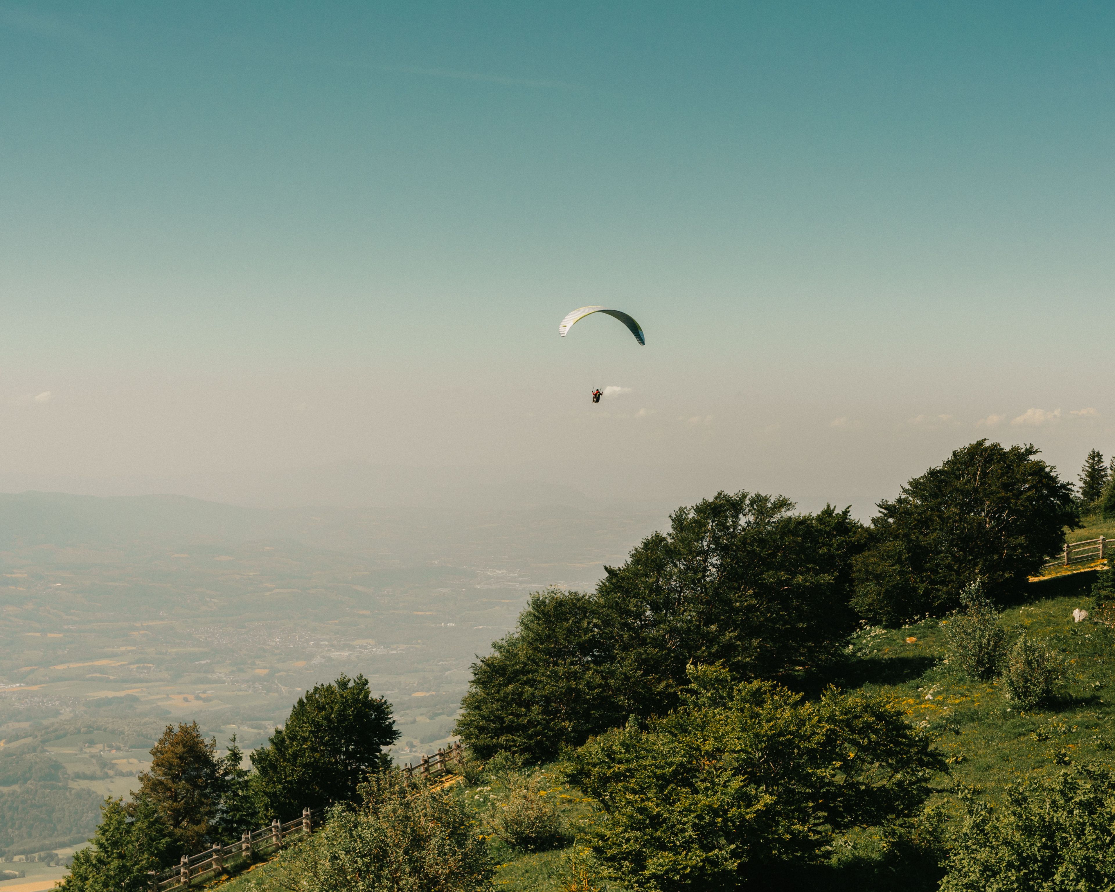 Een persoon paraglidet boven een landschap met bomen en een verre ligging van een stad.
