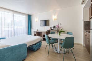 Habitación de hotel con cama, escritorio, mesa de comedor con sillas, televisor montado en la pared y zona de cocina.