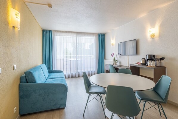 A modern hotel room with a sofa, dining table, chairs, a flat-screen TV, and a kitchenette.