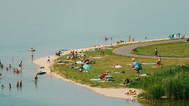 Zona junto al lago con gente nadando y tomando el sol, un área de césped con tiendas de campaña y un bote en el agua.