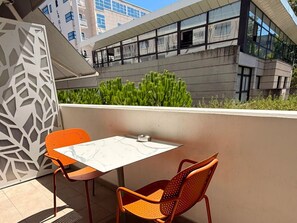 A balcony with two chairs and a table, overlooking a building with large windows and greenery.