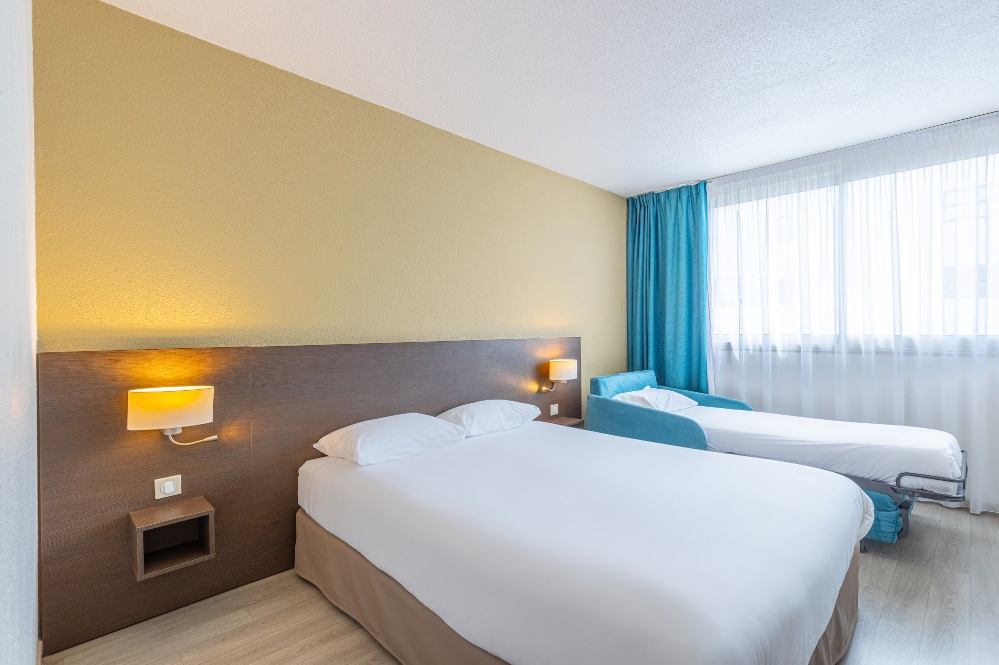 Une chambre d’hôtel avec deux lits, une table de chevet, une lampe fixée au mur et une fenêtre avec des rideaux.
