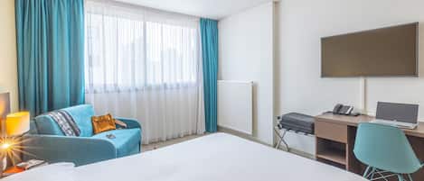 A hotel room with a bed, a desk with a computer, a chair, a TV, and a window with curtains.