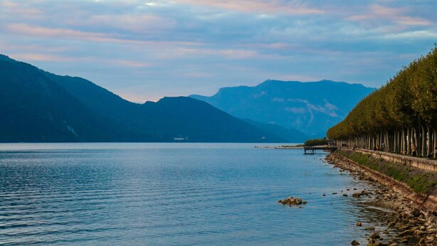 Un lago tranquillo con montagne sullo sfondo e un sentiero alberato lungo la riva.