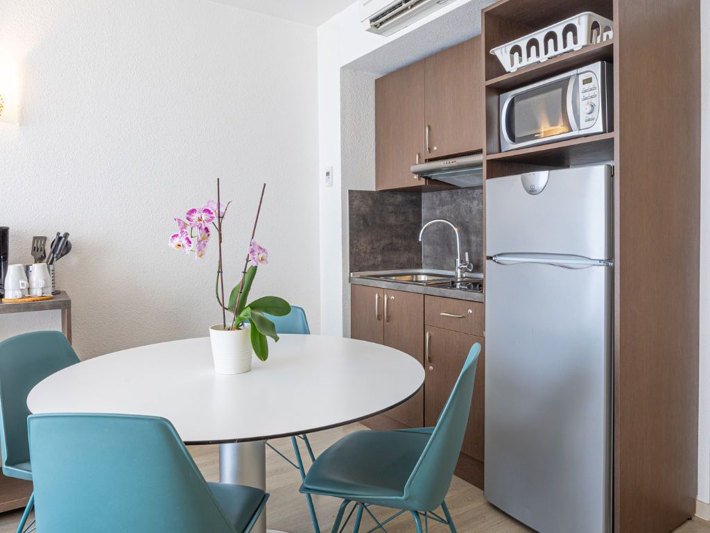 Une cuisine moderne équipée d’un réfrigérateur, d’un four à micro-ondes et d’un évier en acier inoxydable. On y trouve une table ronde avec des chaises et une plante en pot, une orchidée.