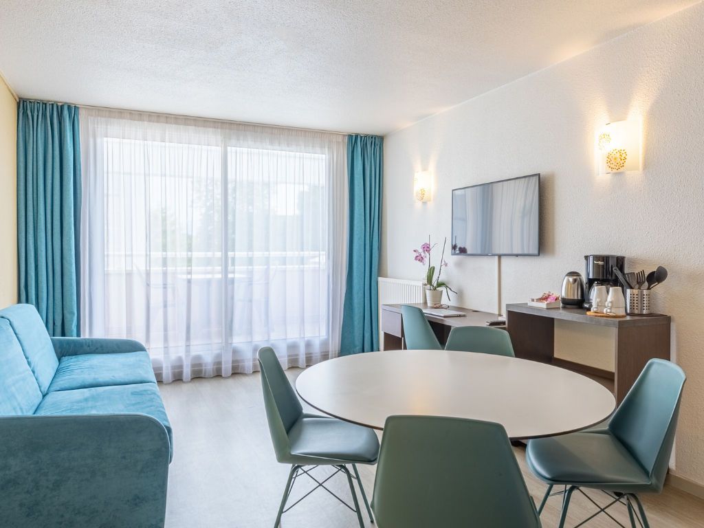 Quarto de hotel com uma mesa de jantar redonda, cadeiras azuis, um sofá, uma TV fixada na parede e uma janela com cortinas.