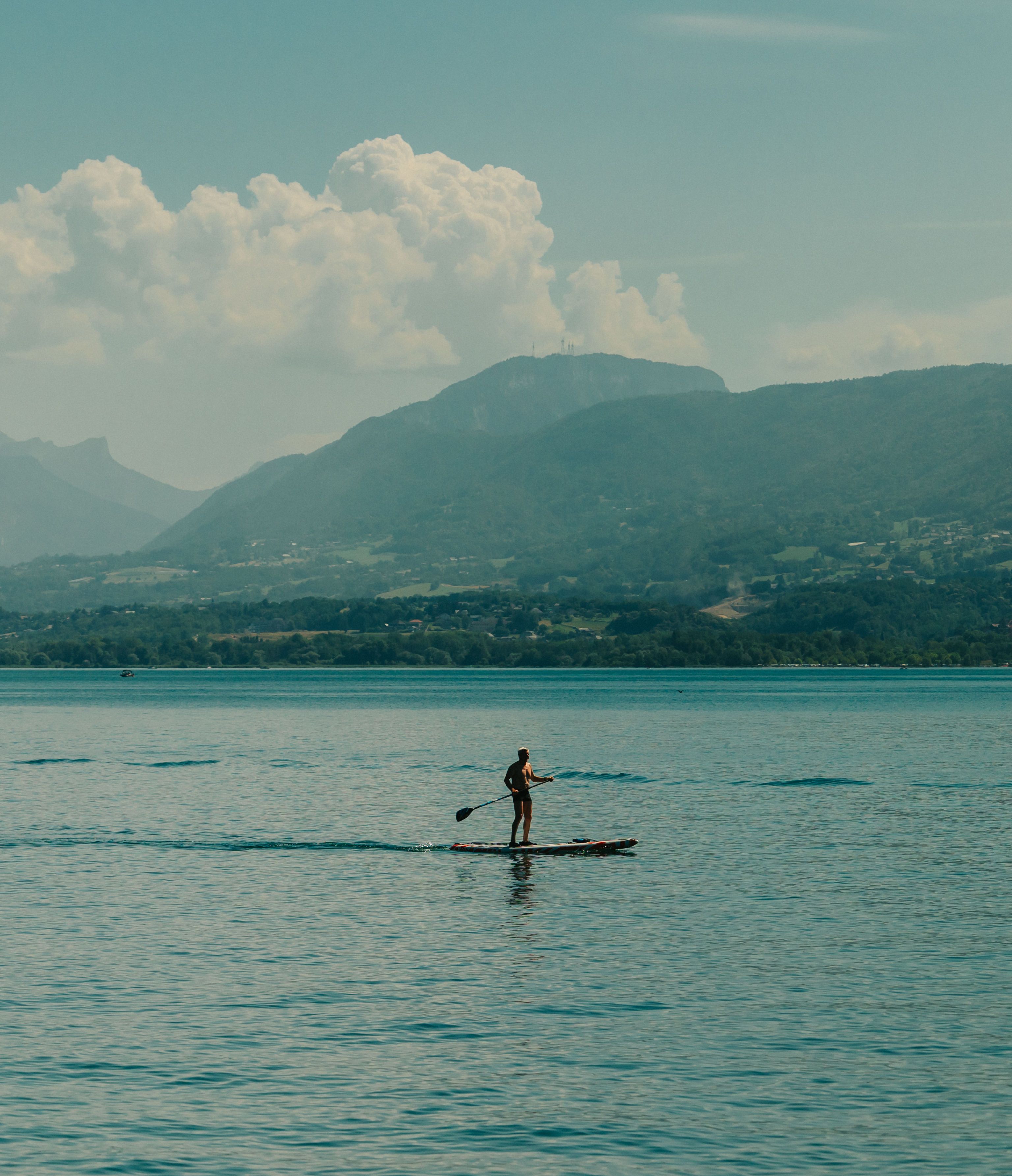 En person står på paddleboard på et roligt vandareal med bjerge i baggrunden.