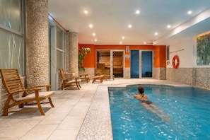 Una piscina interna con una persona che nuota, circondata da sedie di legno e un muro in pietra.
