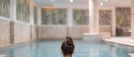 A person sitting at the edge of an indoor swimming pool with a tiled floor and a view of the pool's interior.