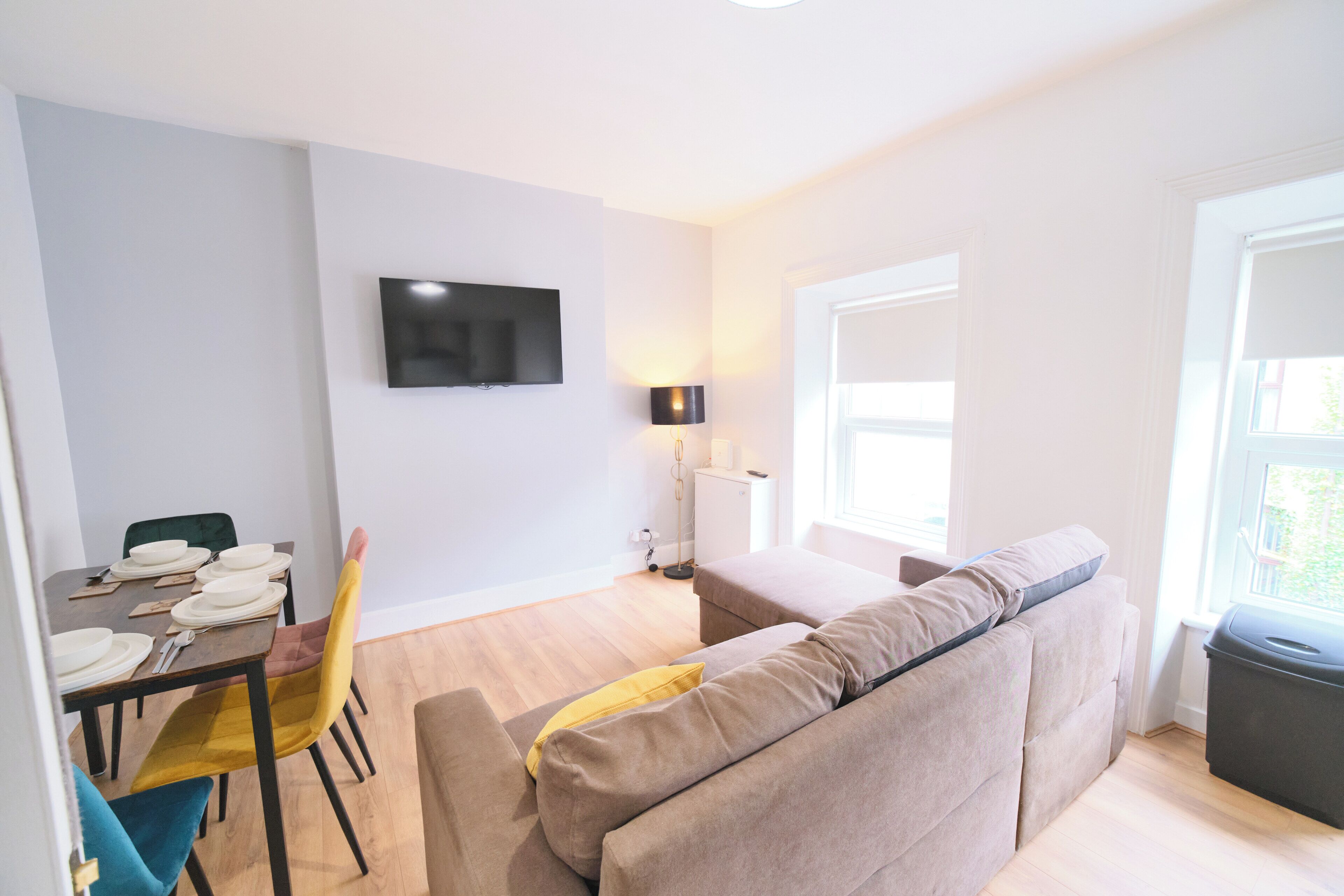 A modern living room with a sofa, a dining table set for two, a flat-screen TV mounted on the wall, and a window with a view.