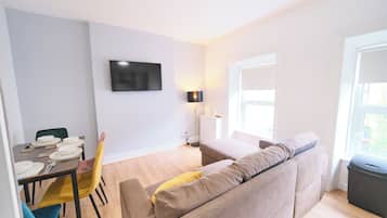 A modern living room with a sofa, a dining table set for two, a flat-screen TV mounted on the wall, and a window with a view.